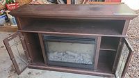 Wide view of dark brown wood electric fireplace cabinet with glass paneled doors open, showing faux logs inside fireplace insert.