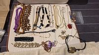 Top-down view of the diverse necklace collection laid out on a beige surface including beaded, pendant, and chain necklaces in purple, wood-tone, gold, black, and pearl colors.