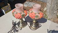 Two pink floral glass shade table lamps with brass bases and power cords, shown side by side on an outdoor table.