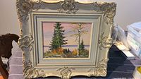 Front view of framed wood painting with decorative cream colored frame featuring pine trees and autumn landscape with sunset sky.