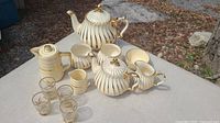 Complete set displayed outdoors showing teapot, sugar bowl, creamer jug, two cups, and six glass shot glasses with gold pattern.