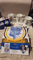 Overview of five commemorative items including mugs, salt and pepper shakers, tea towel in packaging, arranged on beige cloth