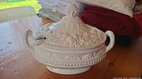 White stoneware casserole dish with matching lid on wooden surface, showing embossed fruit and leaf design on lid and dish with handles