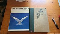 Two bird books on a wooden table, one with dust jacket showing a bird in flight on blue background, titled The Birds of Canada.