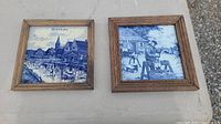 Two framed blue Delft ceramic tiles, side by side on a surface, showing different rural blue and white scenes