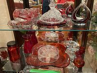 Overall view of vintage art glass collection on glass shelving showing jars, dishes, ashtray, bottles, and candle holders in clear, red, and amber glass.