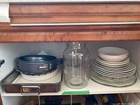 Shelf with two springform pans, ceramic pie plate, large glass jar with no lid, stacked ceramic plates and bowls, and metal baking tray with handle.