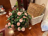 Photo shows a large bunch of artificial pink rose flowers with green leaves and two woven baskets, one natural tan round basket with handles and one smaller white rectangular basket.