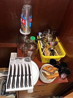 Photo showing disposable glasses stacked on glass jar, plates, coasters, black mugs, and a yellow plastic basket with kitchen utensils including ladle and brushes, and a metal pot with steamer insert and colander