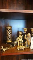 Photo showing all three brass items on a wooden shelf: cylindrical candle holder, ornate easel, bird-shaped clamp/scissors
