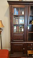 Front view of the tall wood cabinet showing glass door with shelves inside holding books and other items, and lower wooden door section.