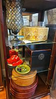 Shelf showing a large silver and black mosaic candle holder on black pedestal, a large gold and white mosaic candle holder behind it, a black wood box with latch, stacked containers below, a woven basket with lid, red fabric hearts, and a small green matchbook.