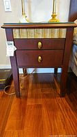 Front view of wooden side table showing two drawers with brass oval pull handles, slatted wood decorative panels under glass top.