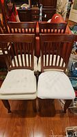 Four wood framed chairs with vertical slat backs in medium brown finish, beige upholstered seats showing stains and wear, arranged in two rows of two on a hardwood floor. Several seat cushions included.