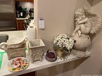 Wide view of lot showing ceramic cherub statuette sitting on a textured spherical base with wings, cream ceramic plant pots with ornamental relief, small faux flower arrangement, purple pin cushion, and hand-painted floral plate.