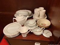 Overall view of china lot containing plates, teapot, creamers, gravy boat, bowls, mugs, butter dish, and nappies arranged on wooden surface.