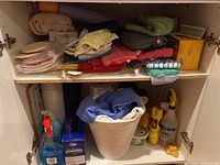Overall view of a cabinet shelf with assorted cleaning cloths folded on top shelf and cleaning liquids including window cleaner bottles and wastebasket with cloths on bottom shelf
