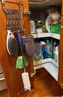 Close-up photo of the inside of a wooden cabinet door showing cleaning brushes and a brown Paderno oven mitt hanging on metal hooks.