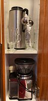 Photo of upper cabinet shelf showing Zojirushi insulated coffee carafe, Cuisinart coffee grinder, and two tall glass coffee mugs with handles along with small coffee accessory brushes