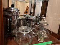 Overview of multiple glass French presses and two glass teapots with small glass mug containing decorative pieces in front