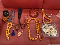 Multiple jewellery pieces displayed on red leather surface: metal pin box with assorted brooches and pins, various beaded necklaces in black, silver, orange and brown, blue tassel necklace, black crochet necklace, shell jewellery components in plastic tub