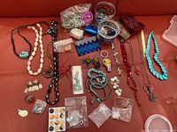 Full overview of costume jewelry lot on red surface including necklaces, earrings, bracelets, keychains, and purses, some items in small plastic bags.