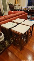 Four wooden counter height stools with cream-colored seat cushions placed together on a hardwood floor next to furniture.