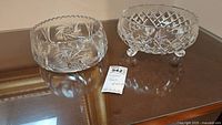 Two clear crystal bowls on a reflective surface, showing one bowl with pinwheel cut and scalloped edge, the other with diamond cut and three ball feet.