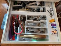 Wide view of drawer with cutlery including forks, spoons, knives, red handled tongs, scissors, timers, and other kitchen tools neatly arranged in white plastic trays.