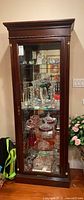 Full front view of tall dark wood display cabinet with glass door and interior glass shelves filled with glassware (contents not included in lot)