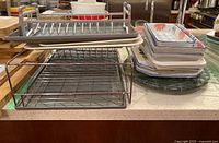 Two metal dish drying racks with plastic drip trays, stack of ceramic baking dishes, greenish marble lazy susan and trivets on a kitchen counter.