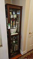 Tall wooden display cabinet with glass front door, multiple glass shelves inside, containing various decorative items. Wood is dark brown with some light chips and wear.