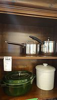 Two stainless steel pots with lids on wood shelf above green Staub cocotte and white canister