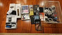 Lot of assorted electronics on hardwood floor showing multiple boxed and loose items including phone cases, cables, adapters, and headphones.
