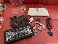 Photo showing various evening bags including a white beaded clutch with rhinestones, a dark beaded bag with blue accents, and a black rectangular beaded clutch, alongside assorted brooches and pendants laid out on a red surface.