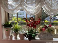 Window sill arrangement of six faux plants and planters including green ferns, red orchids, and white floral planter.
