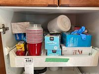 Kitchen cabinet shelf with stacked red and clear disposable cups, boxes of freezer bags and other storage bags, blue box of gloves, and garbage bag rolls.