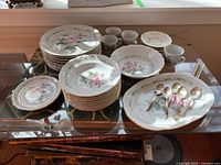 Wide shot showing the whole dinnerware set arranged on a transparent glass table, including stacked dinner plates, smaller plates, bowls, cups, saucers, large serving platter with teaspoons on top.
