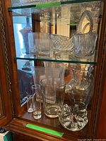 Glass vases, basket, sled-shaped candy dish, and tealight holder displayed on glass shelves inside wooden cabinet.