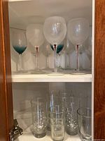 Top shelf of cabinet showing 6 blue-tinted wine glasses with large bowls and long stems, and bottom shelf with 4 clear highball glasses with polka dots and 8 smaller juice glasses with white star or snowflake patterns.