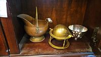 Three metal items displayed in a wooden compartment: a brass coal scuttle on the left, brass globe with stand in the middle, and silver-tone metal bowl with frog decoration on right.