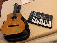 Dixon acoustic guitar resting on a case and Akai MPK25 keyboard lying beside it on a bed.