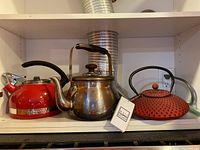 Front view of three metal kettles on shelf: red KitchenAid, stainless steel Well Equipped, red cast iron tea pot
