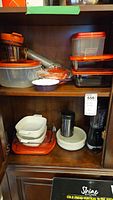 Shelf showing plastic food storage containers with orange lids, glass dishes, Corning Ware casserole dishes white with blue floral pattern, Hamilton Beach blender, and Thermos food container.