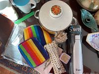 View of ceramic casserole dish, S'well water bottle in box, rainbow-striped dishcloths, brush, wood cutlery set in fabric holder, silver scrubber pad, and small container of seeds or grains on tabletop.