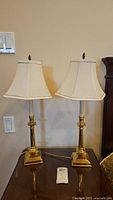 Two matching brass tone metal table lamps with off-white fabric shades on a wooden surface.