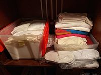 View of two plastic storage bins containing folded white linens, colorful towels, and packaged white slippers on a shelf.