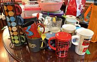 Mixed collection of mugs with colorful and patterned designs, some stacked, set on a cluttered table.