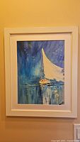 Overall view of framed abstract sailboat painting with blue and white color scheme hung on beige wall.