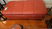 Full view of rectangular reddish-brown leather ottoman bench with stitched panel design on top, positioned on wood floor.